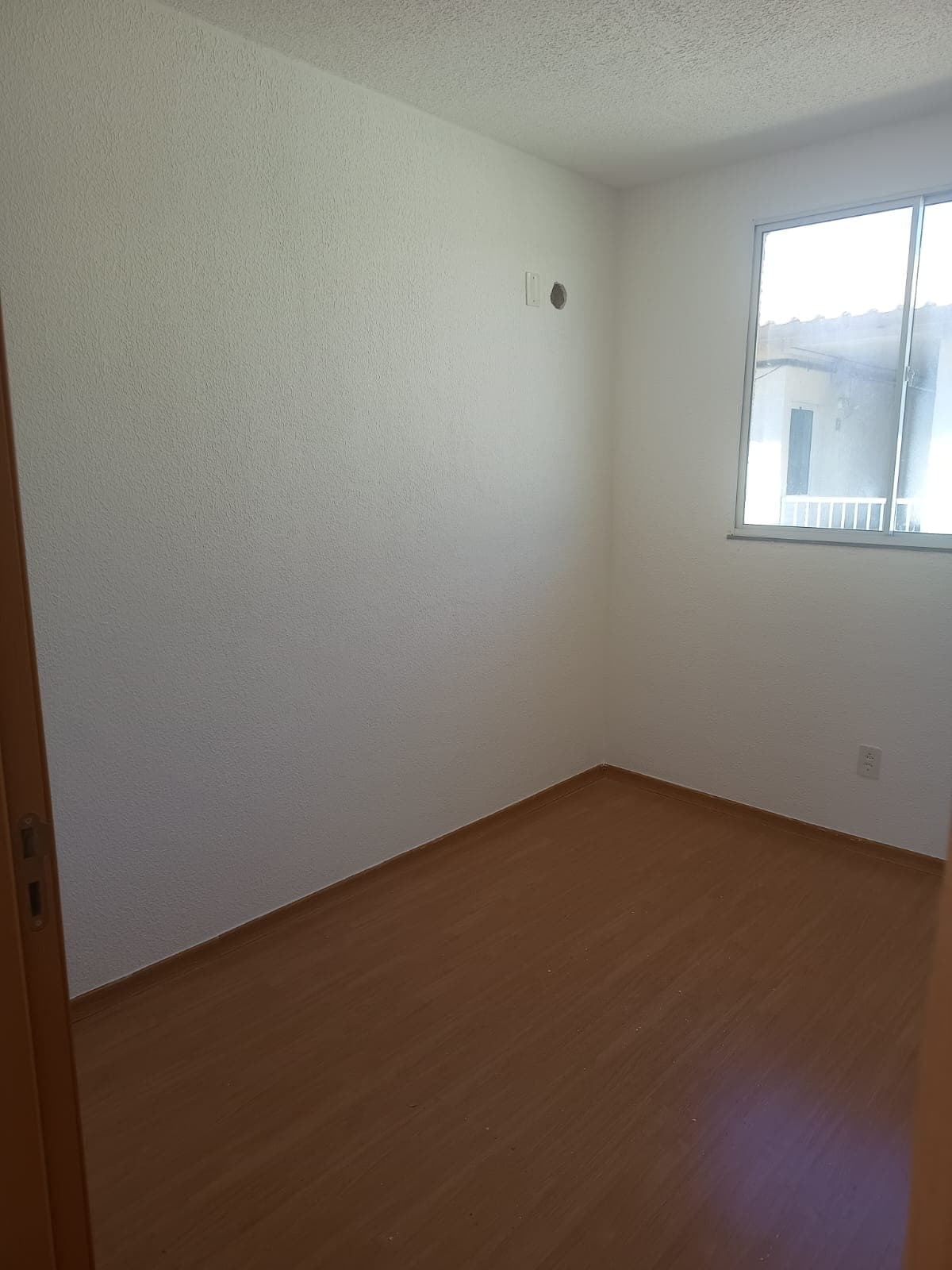Quarto com paredes brancas, piso laminado amadeirado e janela de alumínio.